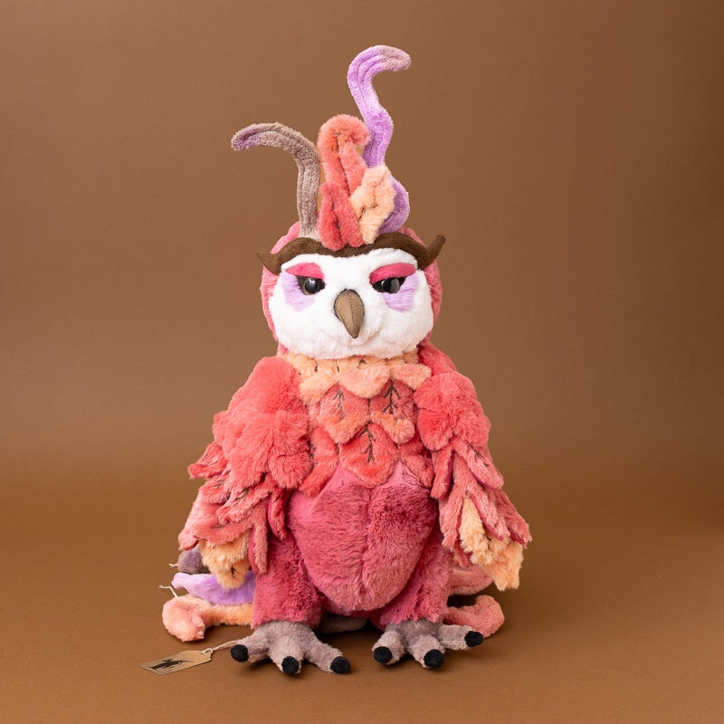 A whimsical, fuzzy jinzi-the-rising-phoenix plush toy styled as a female fairy with pink and orange feathers, a purple horn, and gray claws, set against a solid brown studio background.