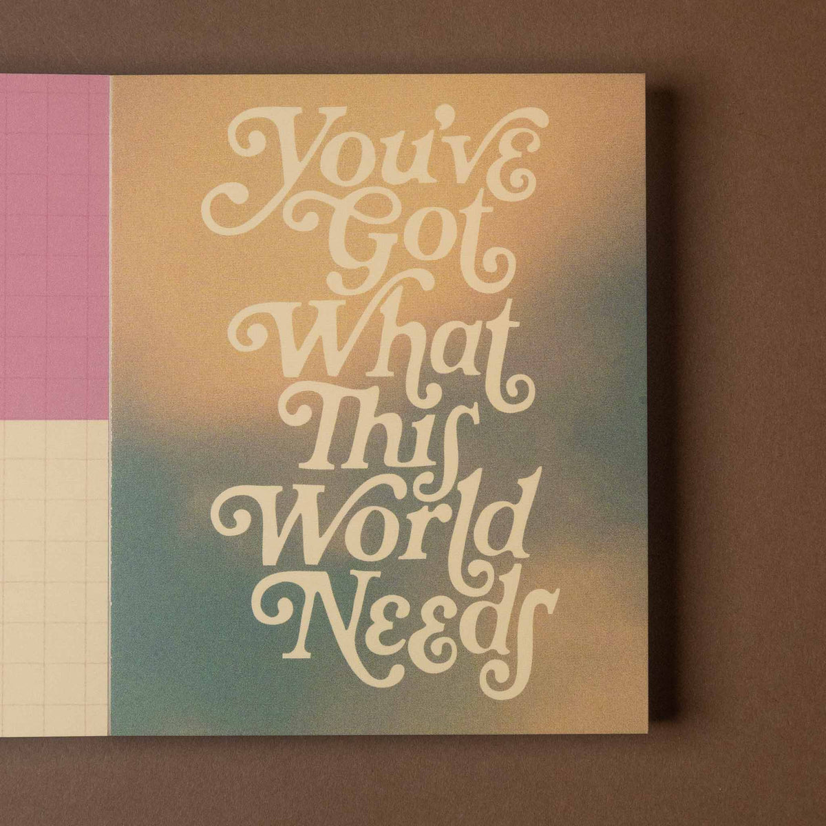 A square art card with the phrase 'You've Got What This World Needs' in large cream script over a soft beige-to-teal gradient background, shown standing against a brown wall. A pink and off-white grid-patterned sheet is partially visible at the left edge.