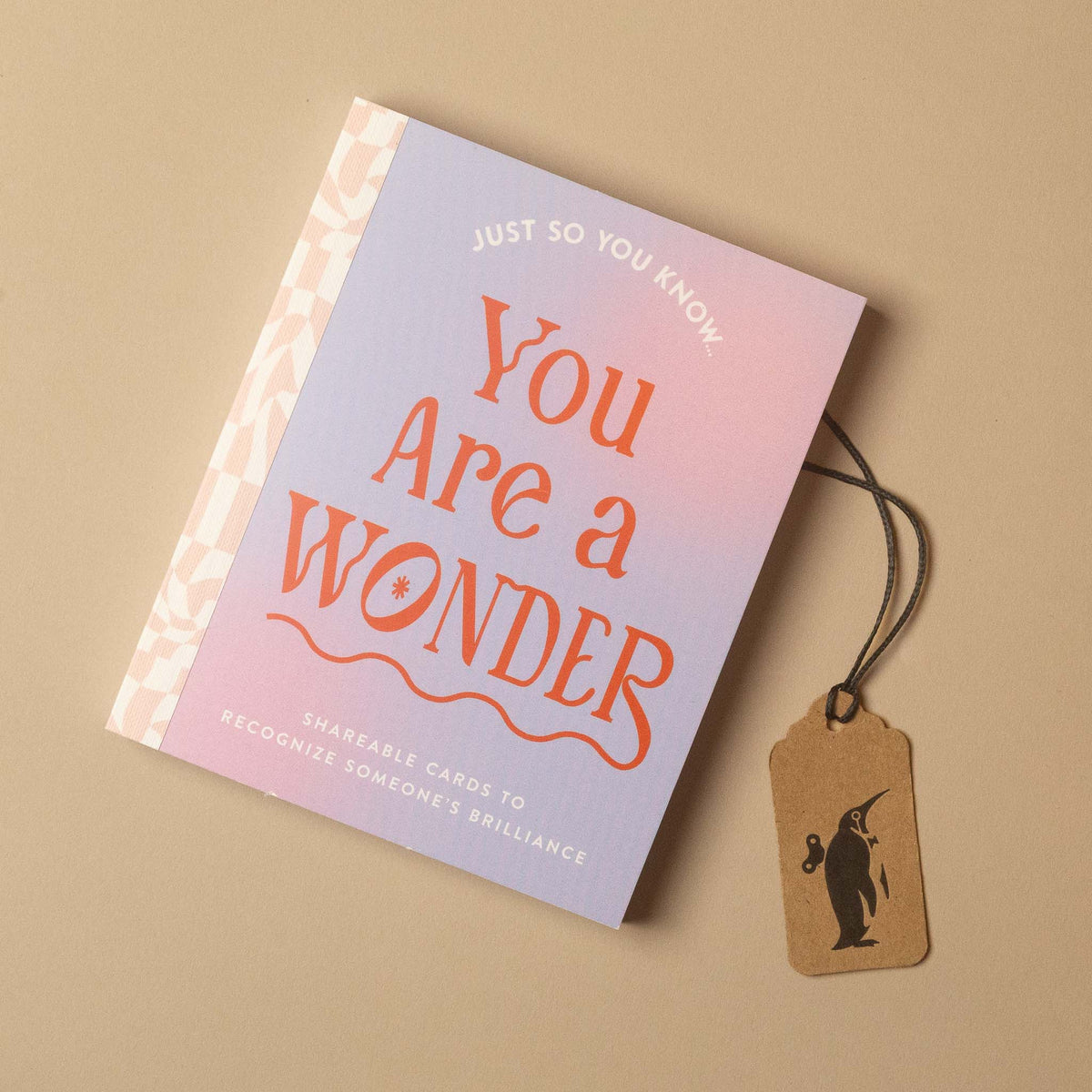 A single greeting card with a pink-to-blue gradient cover reading 'You Are a WONDER' in orange lettering, shown on a beige background with a small kraft paper hang tag attached by a black string.