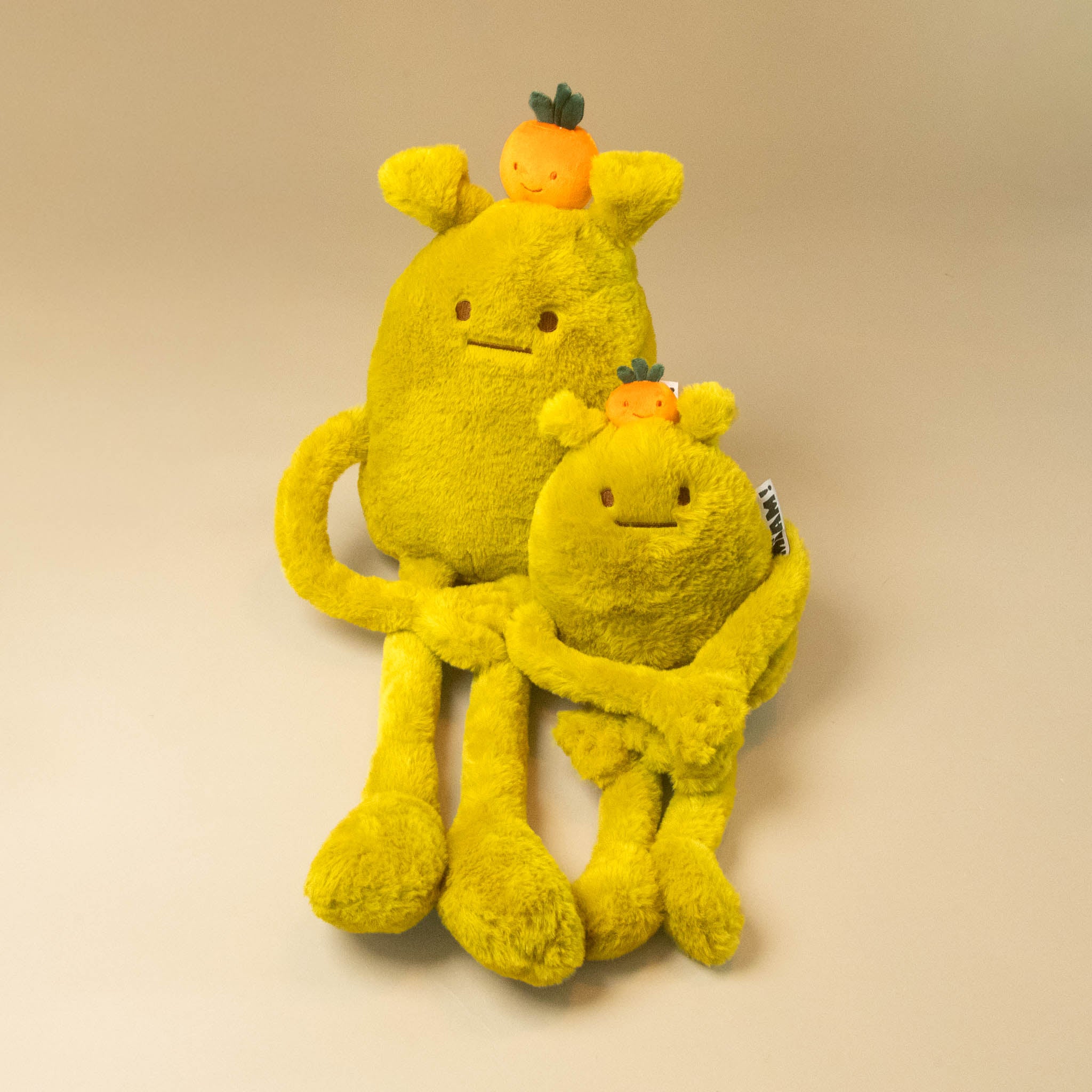 A yellow laurent-jade-grande and petit plush toy with long arms and legs, a small orange fruit on its head on a plain beige studio background.