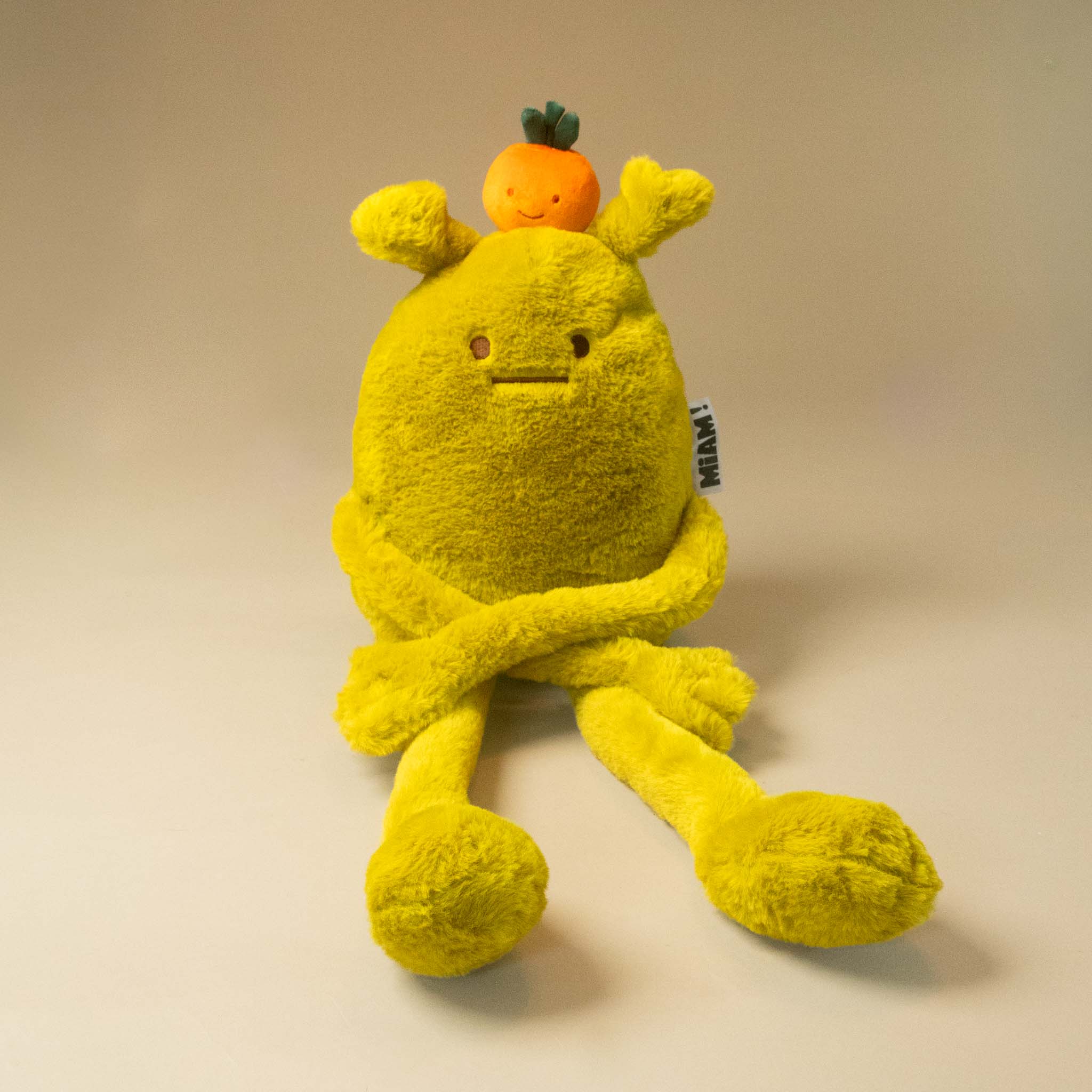 A yellow laurent-jade-grande plush toy with long arms and legs, a small orange fruit on its head on a plain beige studio background.