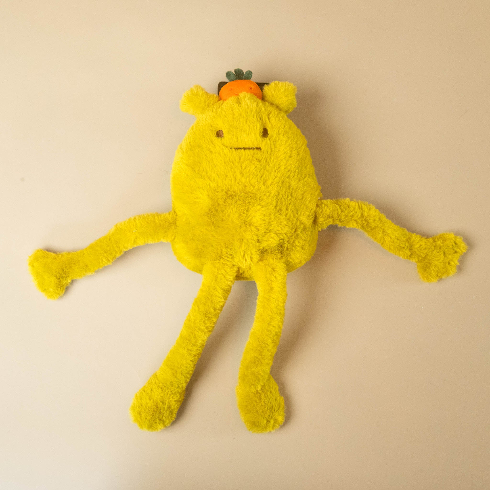 A yellow fuzzy plush toy with long tentacle-like legs and arms, a simple stitched face, and a small orange accent on top, photographed against a plain light beige background.