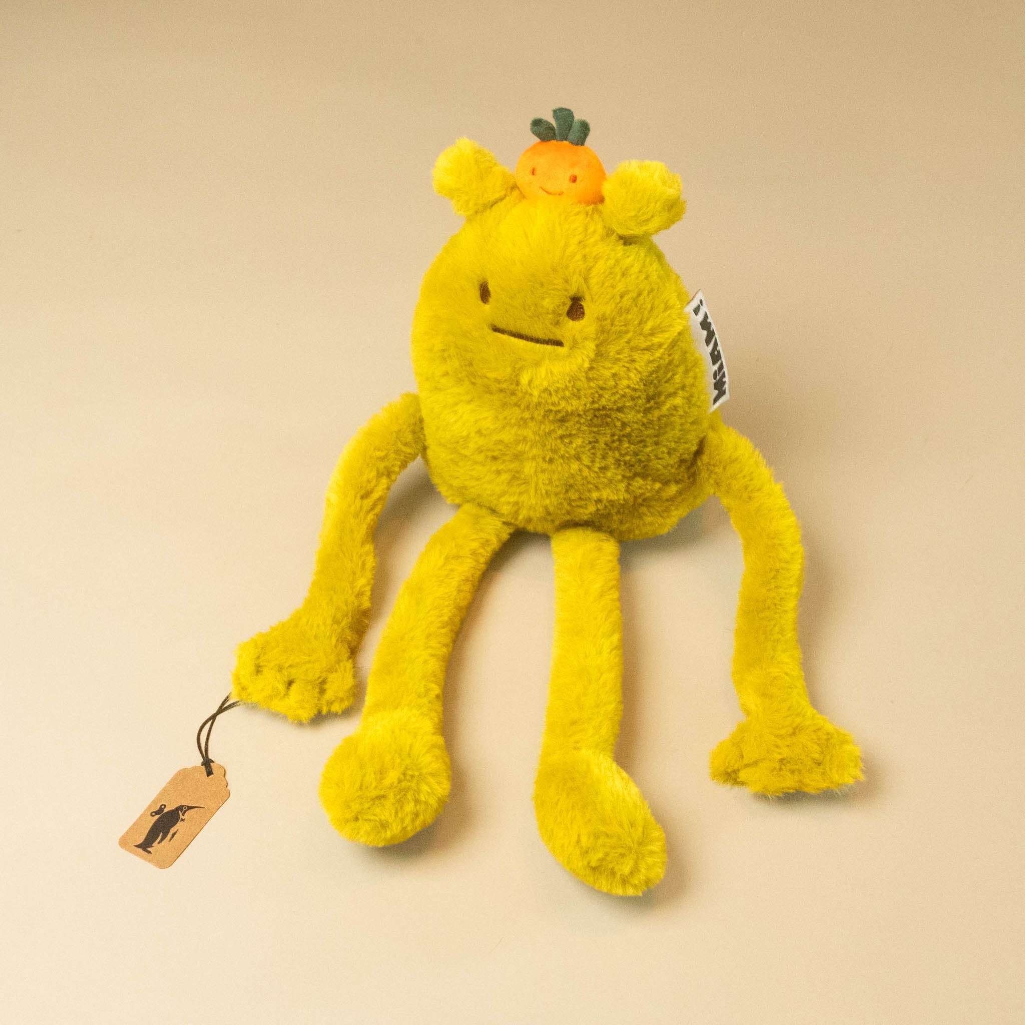 laurent-jade-petit plush toy with a small orange and green jade on its head, standing on a warm beige background. It has long tentacle-like legs and a small stitched smile.