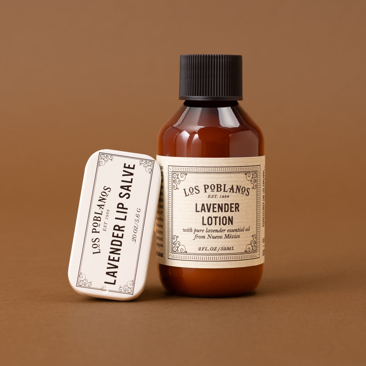 Los Poblanos Lavender Lotion in an amber glass bottle with a black cap, shown next to a white tin of Lavender Lip Salve on a warm brown background.