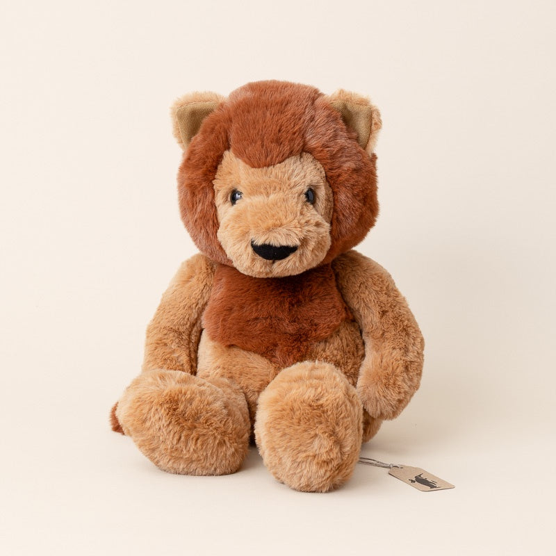 A plush lion toy with a brown mane, tan face and paws, and a small hanging tag, photographed against a plain warm off-white background.