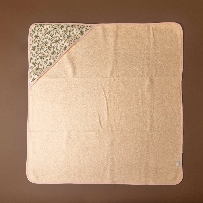 A square baby blanket or towel in pale peach-pink, laid flat on a brown background. One corner is folded over to reveal a floral patterned hood in beige, white, and muted green.
