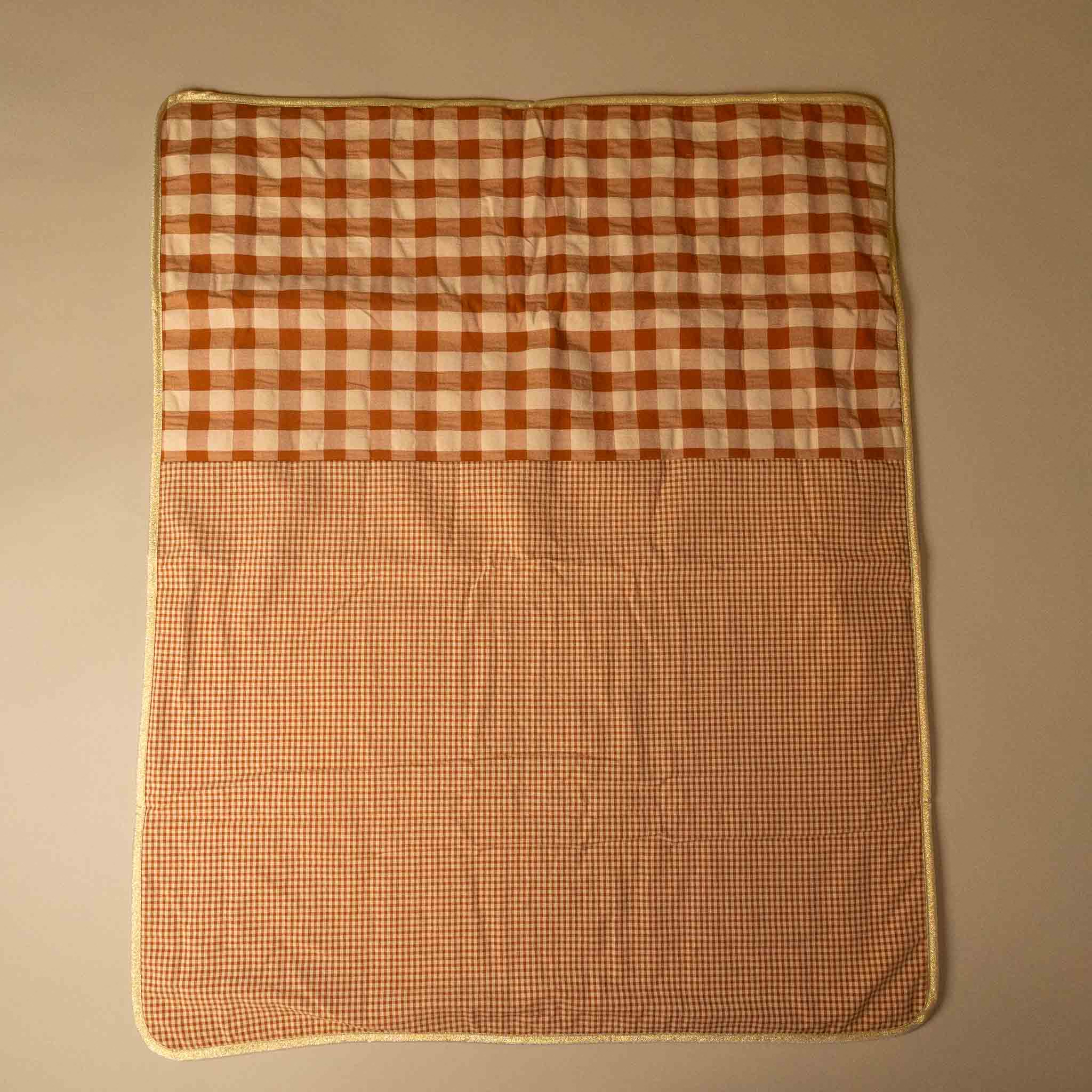 A square quilted blanket or mat with a two-tone gingham pattern, featuring a larger rust-orange and cream checkered top half and a smaller matching checkered bottom half, finished with a cream border, laid flat against a neutral beige background.