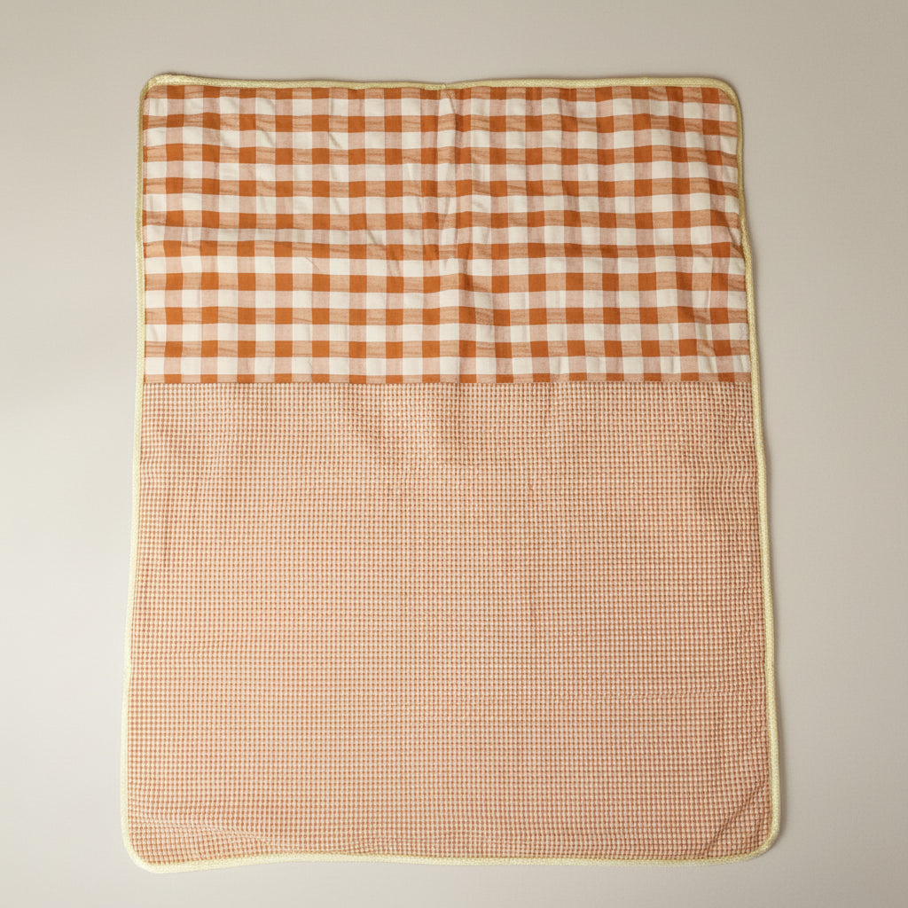 A square quilted blanket or mat with a two-tone gingham pattern, featuring a larger rust-orange and cream checkered top half and a smaller matching checkered bottom half, finished with a cream border, laid flat against a neutral beige background.