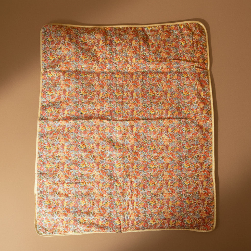A rectangular quilted fabric pad with a dense small floral print in orange, red, yellow, and blue on a light background, finished with a beige border, laid flat on a beige surface.