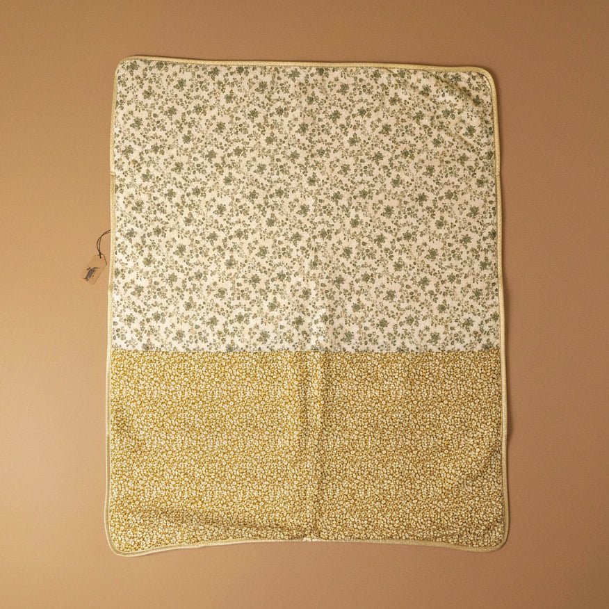 A square fabric blanket or quilt laid flat on a neutral beige background, featuring a two-tone floral pattern. The top half has a small green floral print on an off-white background, and the bottom half has a dense small yellow floral print on a warm beige background.