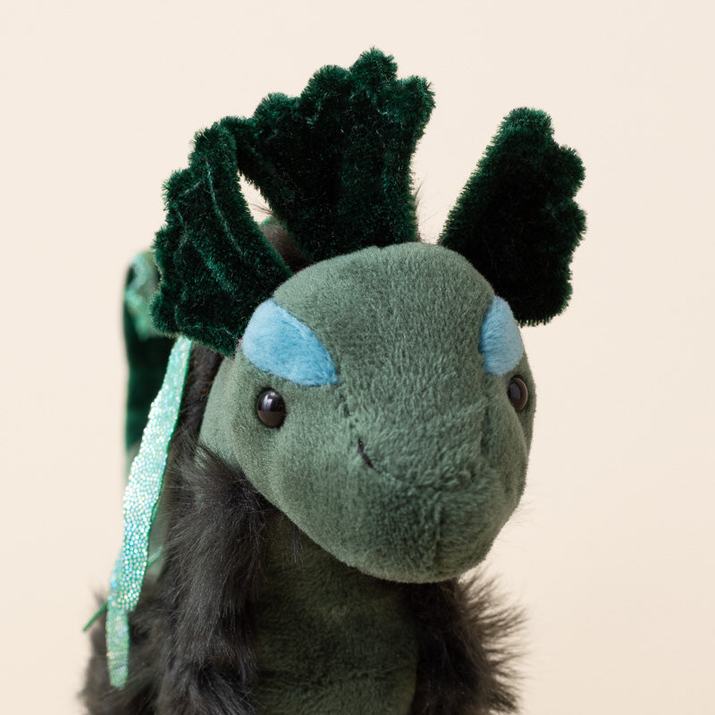 A plush little-nessie-cub toy with a dark green head and back, light blue eye patches, black fluffy underbelly fur, and dark green leaf-like horns, shown against a plain beige background.