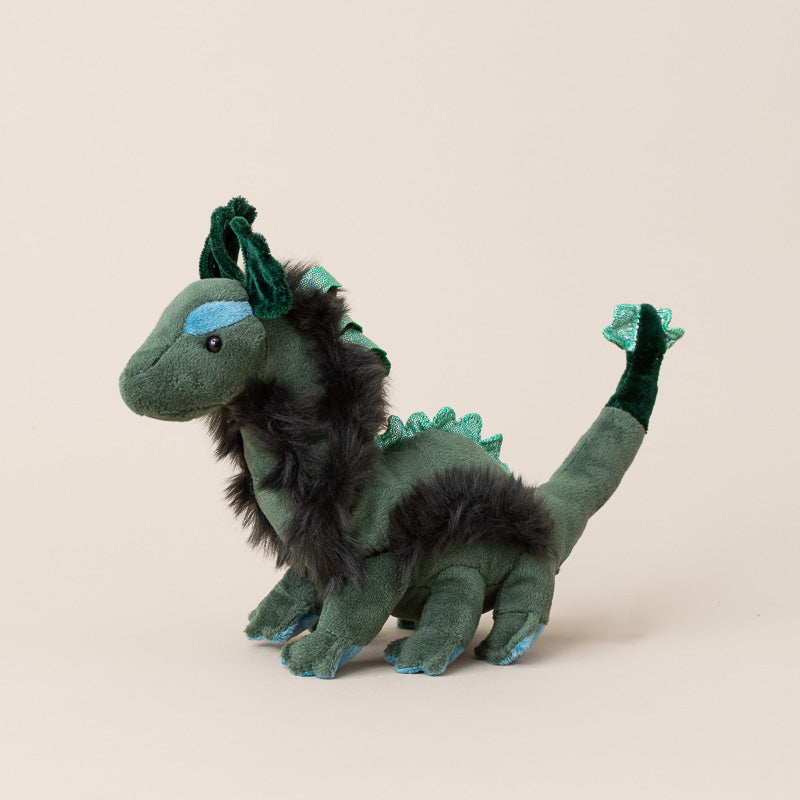 A plush toy little-nessie-cub in dark green velvet with black faux fur accents, blue embroidered eyes and claws, and a glittery green tail, standing against a solid beige background.