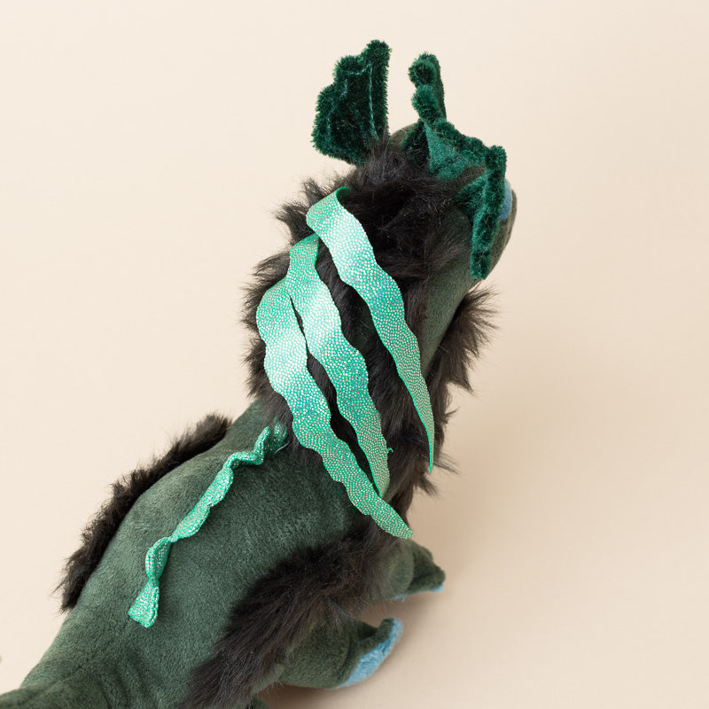 A dark green plush little-nessie-cub toy with black faux fur accents, blue feet, and three glittery mint green claw-like ribbons on its back, shown against a solid beige background.