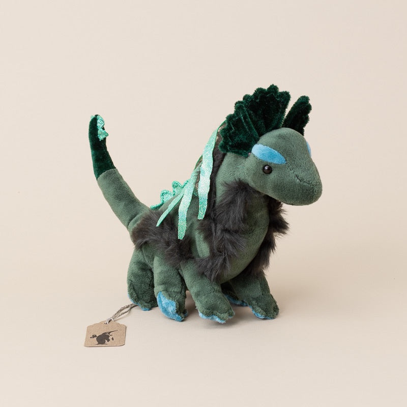 A plush little-nessie-cub toy in shades of dark green, gray, and light blue, standing on a warm beige background with a small hanging tag.