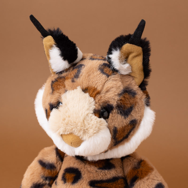 A close-up of a plush lynx toy with brown and black spotted fur, a cream-colored snout and inner ears, and black-tipped ears, set against a solid warm brown background.
