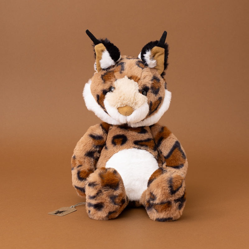 A plush toy lynx with brown and black spotted fur, a cream-colored snout and inner ears, and a white belly, sitting against a solid brown background.