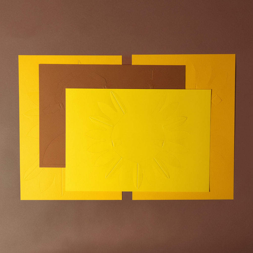 make-your-own-3d-sunflower-paper-model-Three rectangular cardboard pieces in yellow, brown, and orange on a brown background