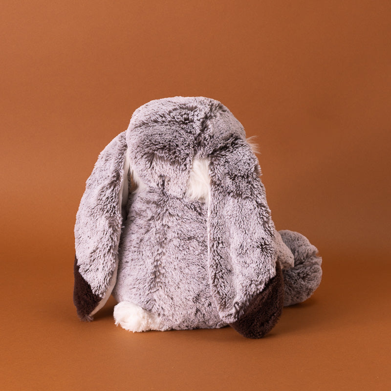 A plush stuffed bunny toy with long floppy ears, sitting upright against a solid warm brown background. The bunny has gray and white fur back- and ears