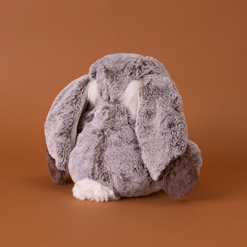 A plush stuffed bunny,seated against a solid warm brown studio background. The toy has soft gray and white fur, dark brown inner ears back