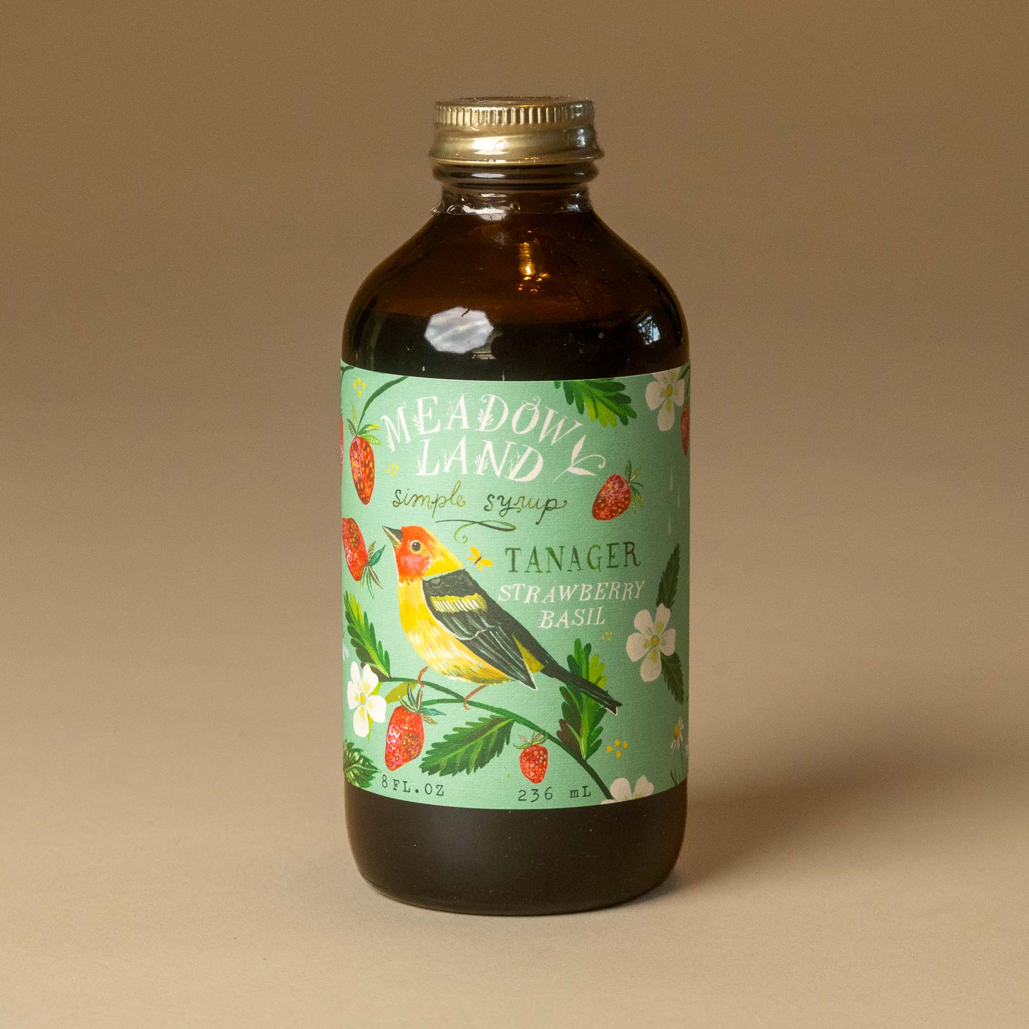 A bottle of meadowland-simple-syrup-tanager in an amber glass bottle with a gold cap and a green illustrated label featuring a yellow and black bird, strawberries, and white flowers, set against a solid beige background.