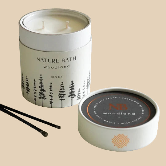 Candle in a metal tin labeled 'Nature Bath Woodland' on a white surface.