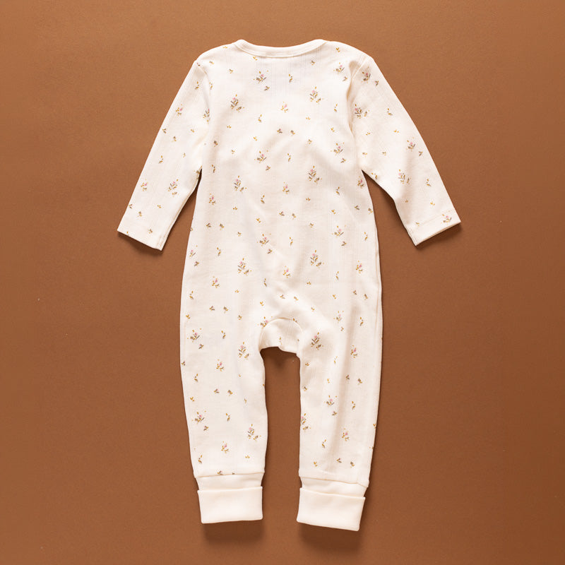 A baby long-sleeve footless jumpsuit in cream white with a small scattered brown floral pattern, laid flat on a solid brown background.