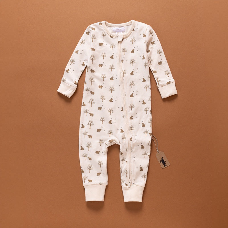 A baby long-sleeve footless one-piece romper in cream with a brown forest animal and tree print, laid flat on a solid brown background with a hangtag attached.