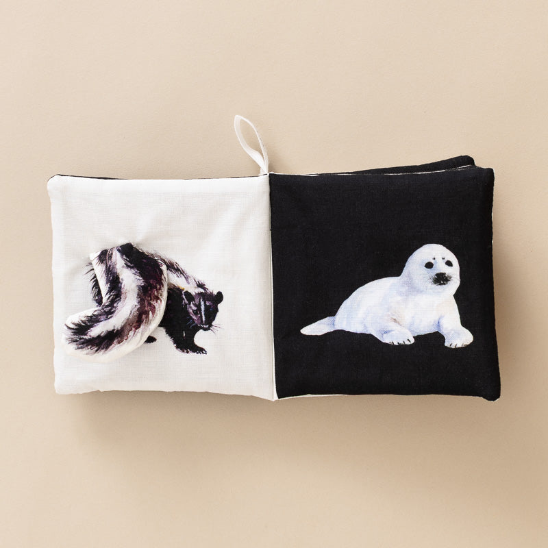 A square fabric book with a split design: one half is off-white with a painted black-and-white skunk, and the other half is black with a painted white seal pupp. The bag has a small hanging loop at the top and is photographed against a plain warm beige background.