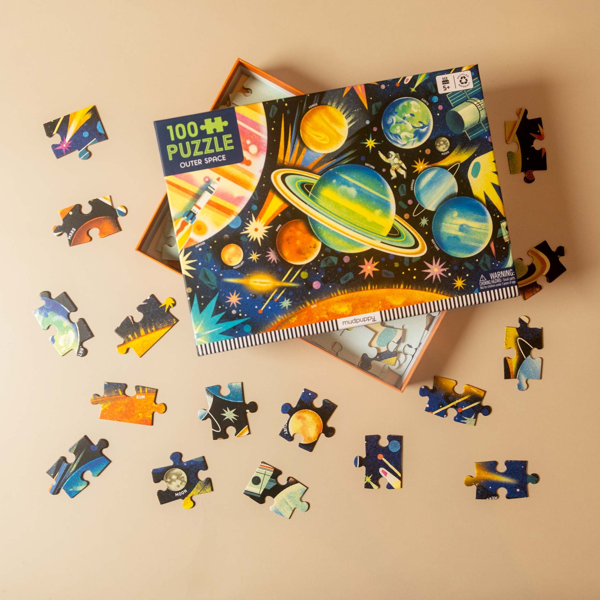 A colorful children's jigsaw puzzle box labeled '100+ PUZZLE OUTER SPACE' is shown on a beige background with several loose puzzle pieces scattered around it. The box artwork features planets, stars, and space-themed illustrations.