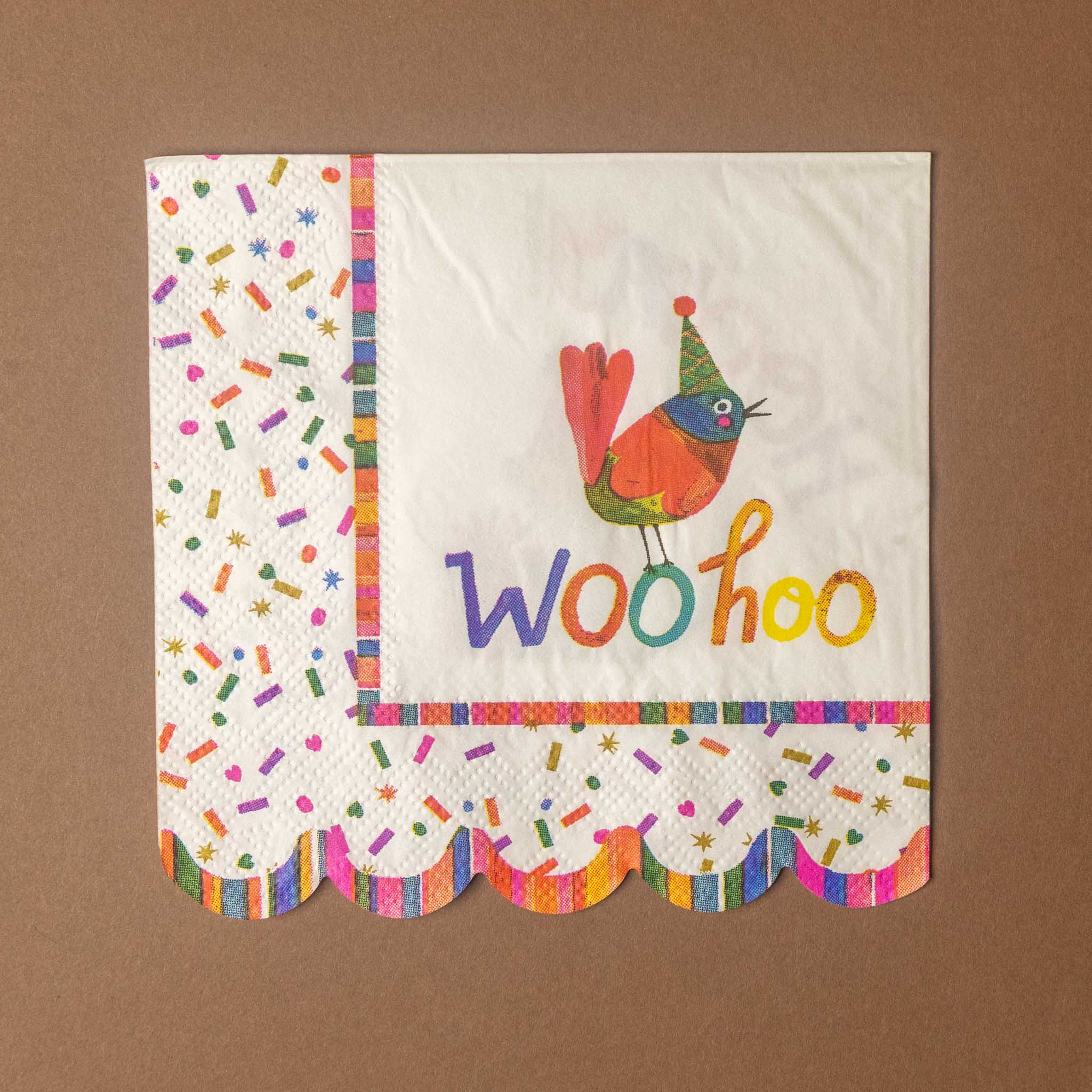 A square white paper napkin with colorful party-themed printing, including a bird wearing a party hat and the word 'Woo-hoo' in multicolored letters, plus sprinkle patterns and scalloped multicolor borders, shown on a brown background.
