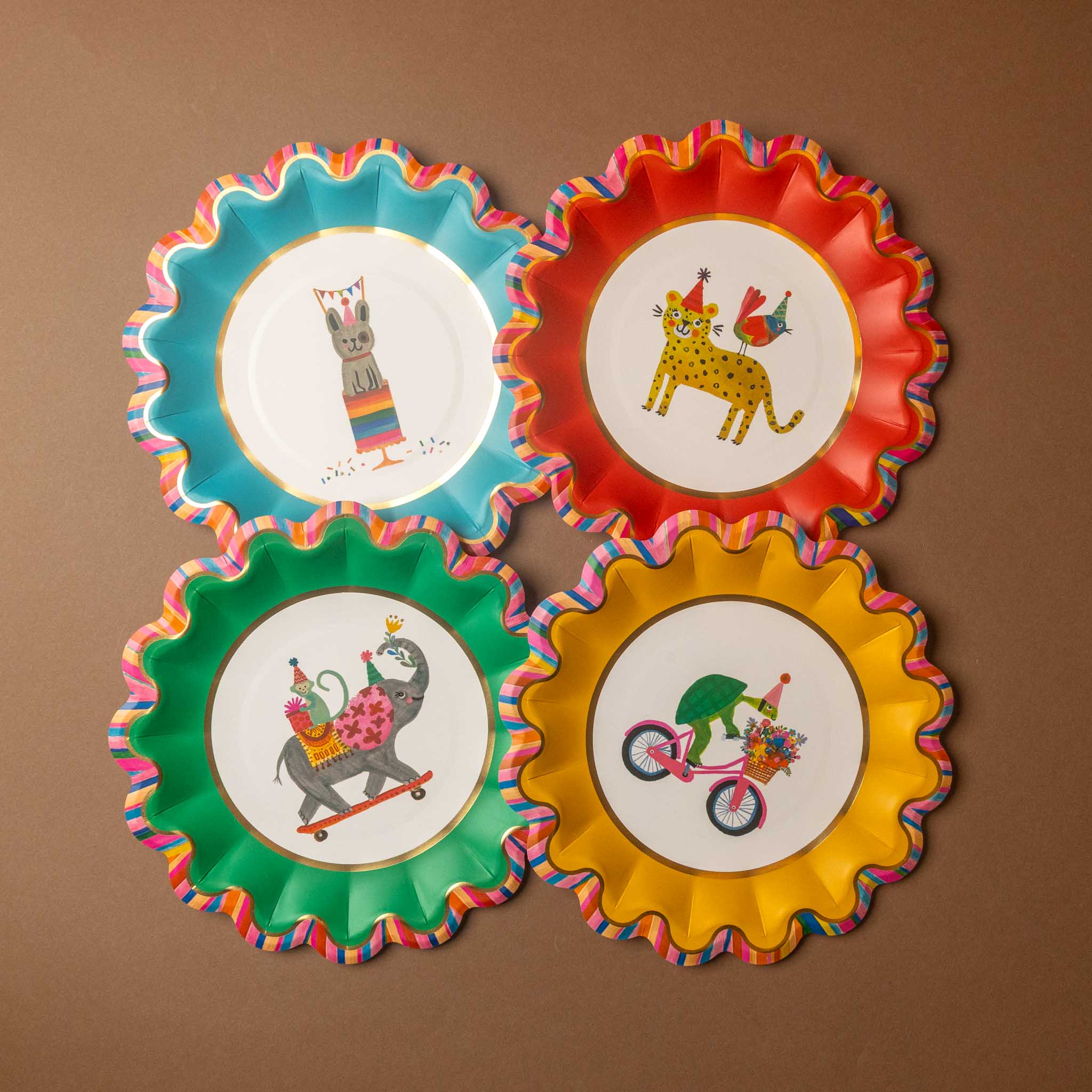 Four colorful party plates arranged in a 2x2 grid on a brown background. Each plate has a scalloped edge with a multicolor striped pattern and a white center featuring a different cartoon animal illustration wearing party hats: a cat, a leopard, a dog on a skateboard, and a bicycle rider.
