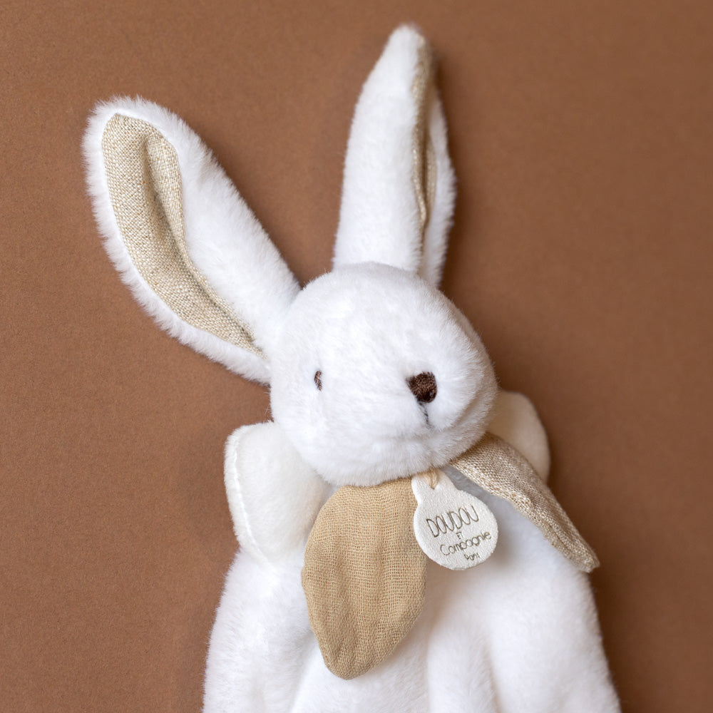 petite-bunny-lovie-happy-wild White plush bunny with beige ears and a tag on a brown background