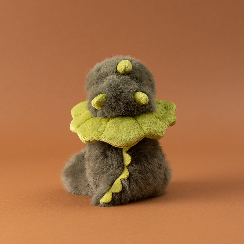 A small plush toy petite-gluepot-dino shown from the back, featuring fluffy gray-brown fur, small yellow horns, and yellow wing-like accents,
