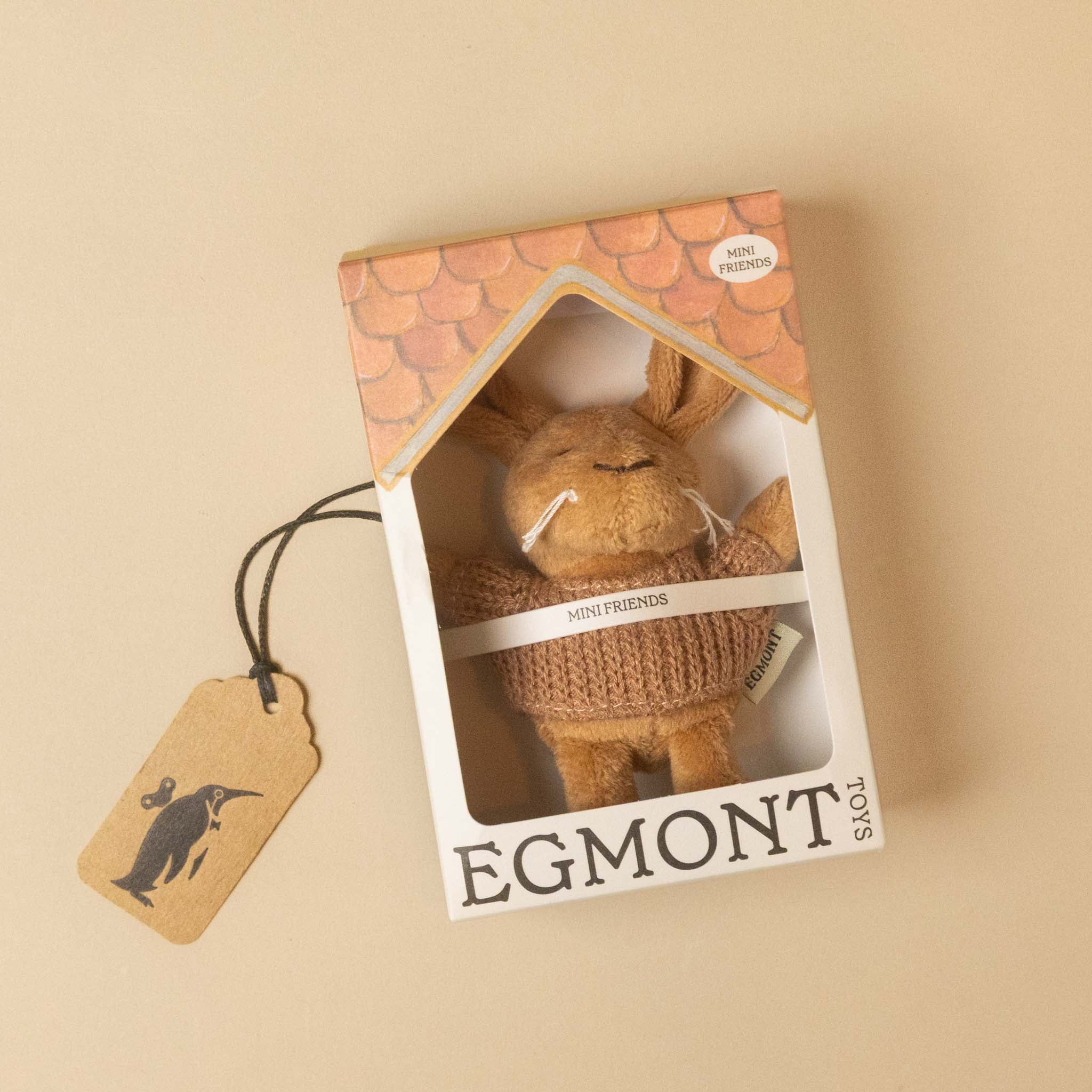 A plush toy petits-amis-lenny-rabbit in a knitted sweater displayed inside its branded house-shaped packaging box, with a hanging paper tag on a warm beige background.