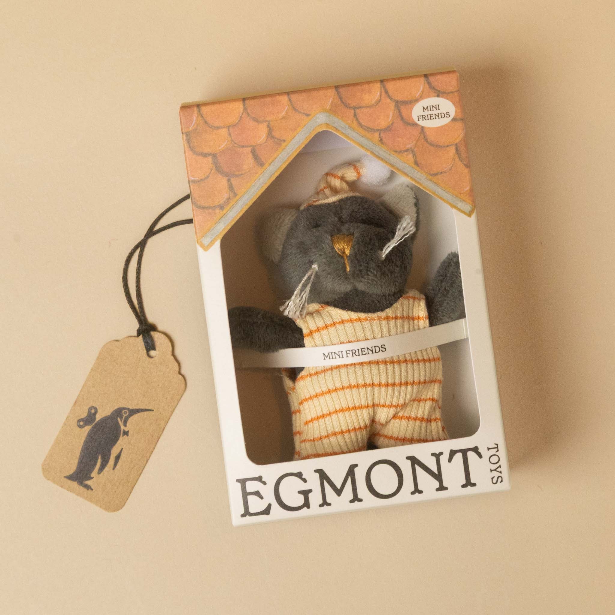A small plush petits-amis-malo-cat inside a decorative window box package, with a hanging kraft paper tag on a warm beige background.