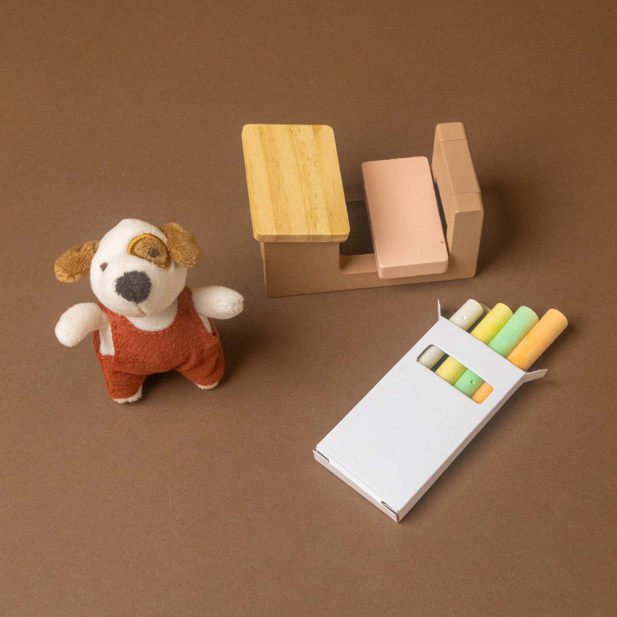 A miniature toy petits-amis-schoolhouse-play-set featuring a small plush puppy in red overalls, a tiny wooden desk and chair, and a white box of pastel chalks with yellow, green, and orange colors, all arranged on a brown background.