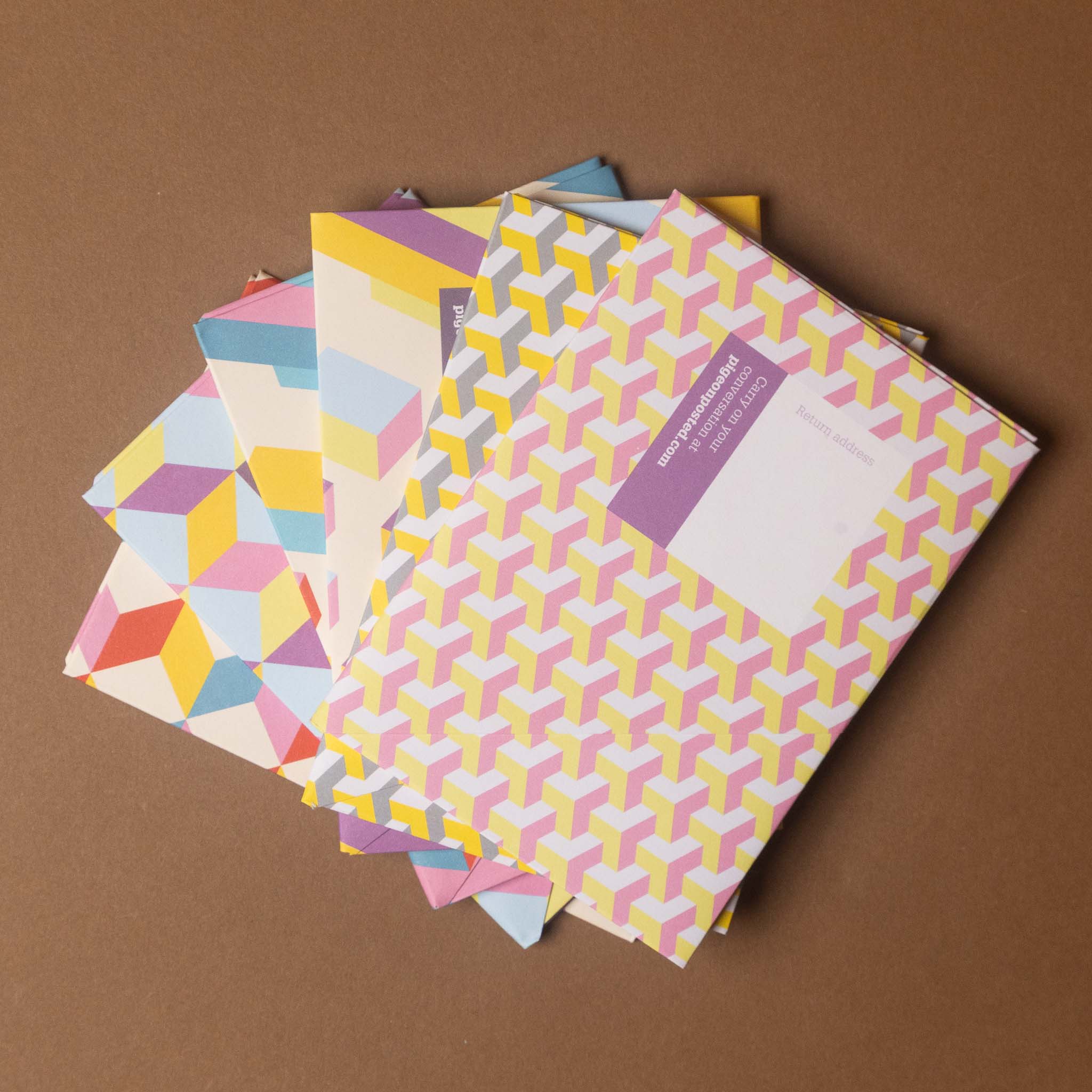 A stack of colorful patterned envelopes or mailers fanned out on a solid brown background. The top envelope features a yellow, pink, and white geometric cube pattern with a purple label, while the envelopes underneath display various pastel geometric designs.