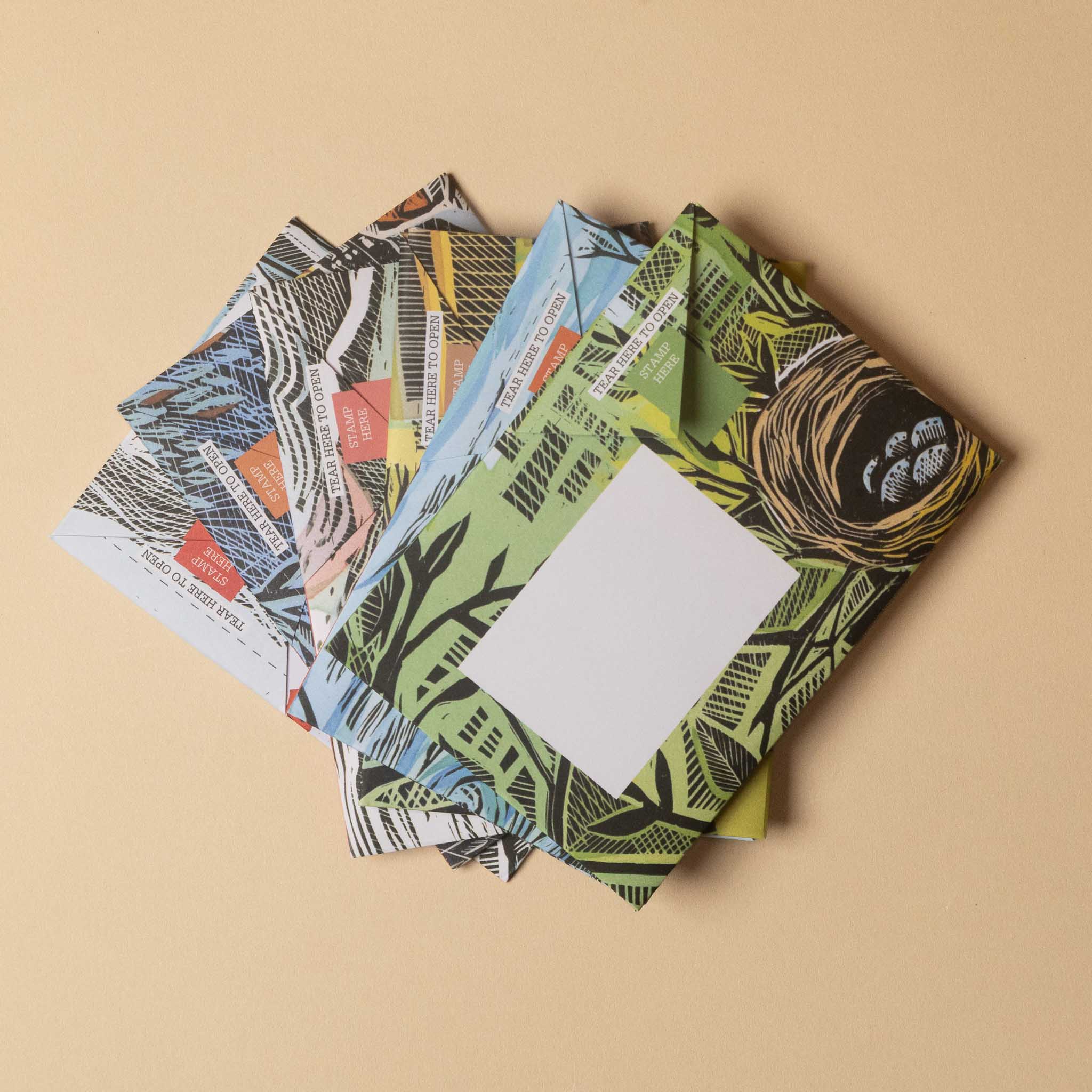 A stack of patterned envelopes or postcards fanned out on a warm beige background. The top card features a green tropical leaf pattern with a white blank center, while the layers beneath show diverse designs including a bird's nest with eggs, a zebra print, and abstract textures.