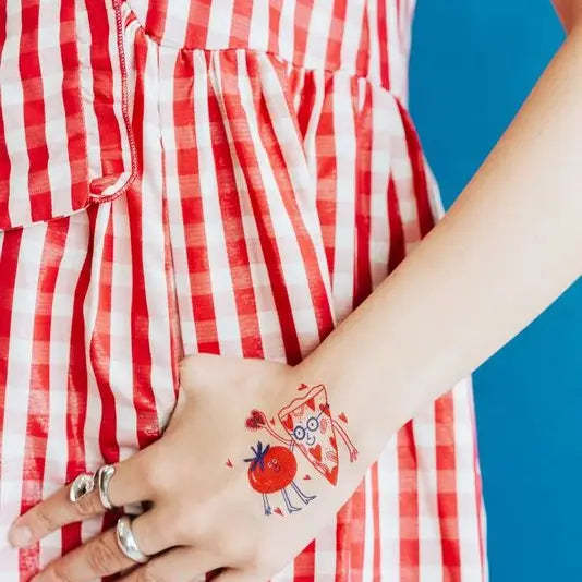 Person wearing a red and white striped dress with a colorful pizza-loves-tomato-temporary-tattoo-pair on their arm against a blue background