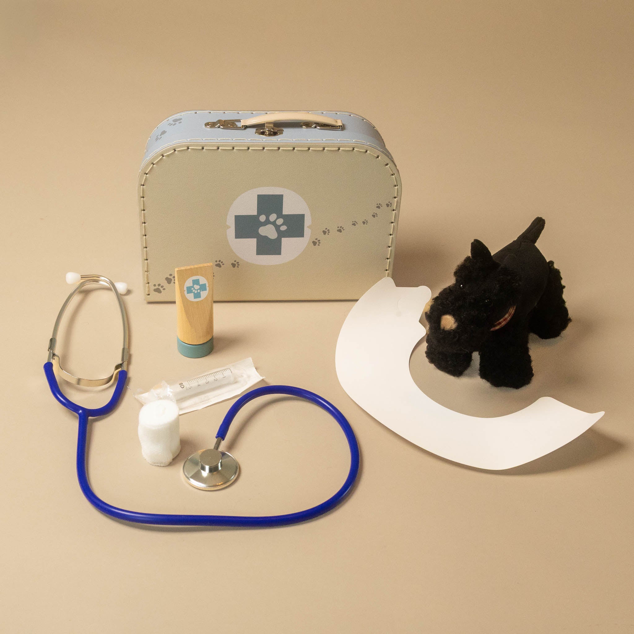 pretend-play-veterinary-case-walter-dog playset arranged on a beige background, including a light blue and cream toy case with a paw-print medical symbol, a blue stethoscope, a small tube, a bandage, a white curved shield, and a black plush dog toy.