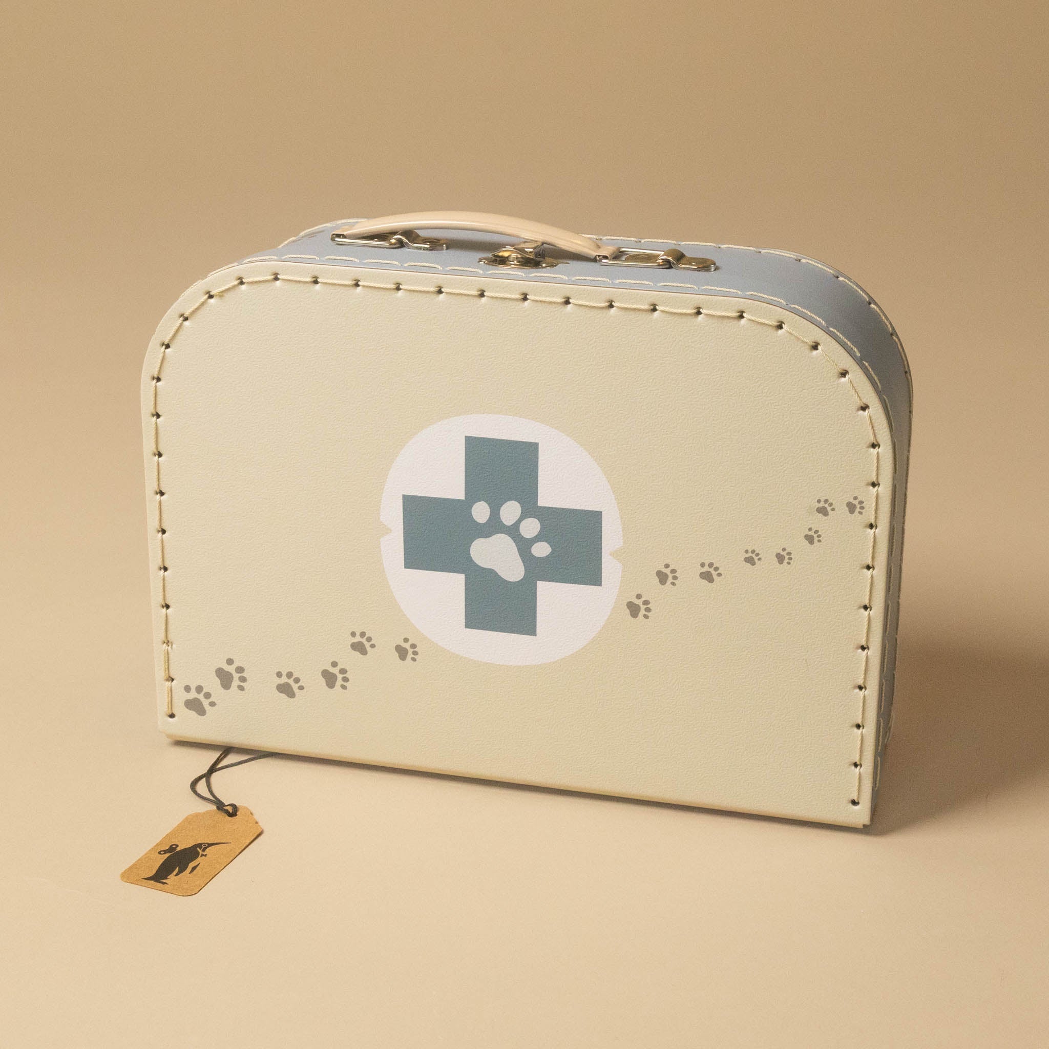 A vintage-style cream-colored suitcase with a blue-gray cross and paw print emblem on the front, featuring decorative stitching, a top handle, and a small hanging tag on a warm beige background.