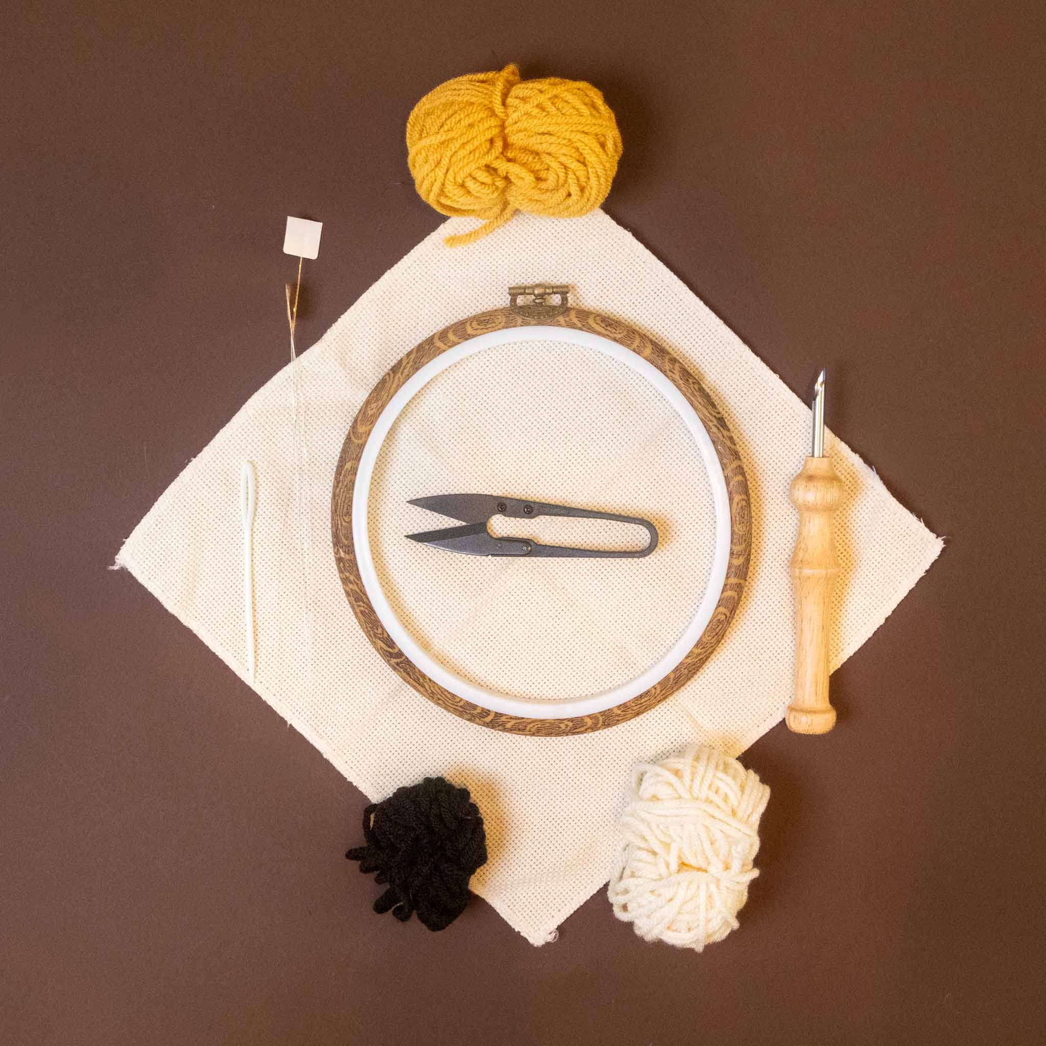 punch-needle-kit-sunshine-y-day tools on a brown background, including a white fabric hoop with a metal needle, a wooden stitch holder, a black thread ball, a cream thread ball, and a yellow thread ball, along with a white square of fabric and a white wooden needle.