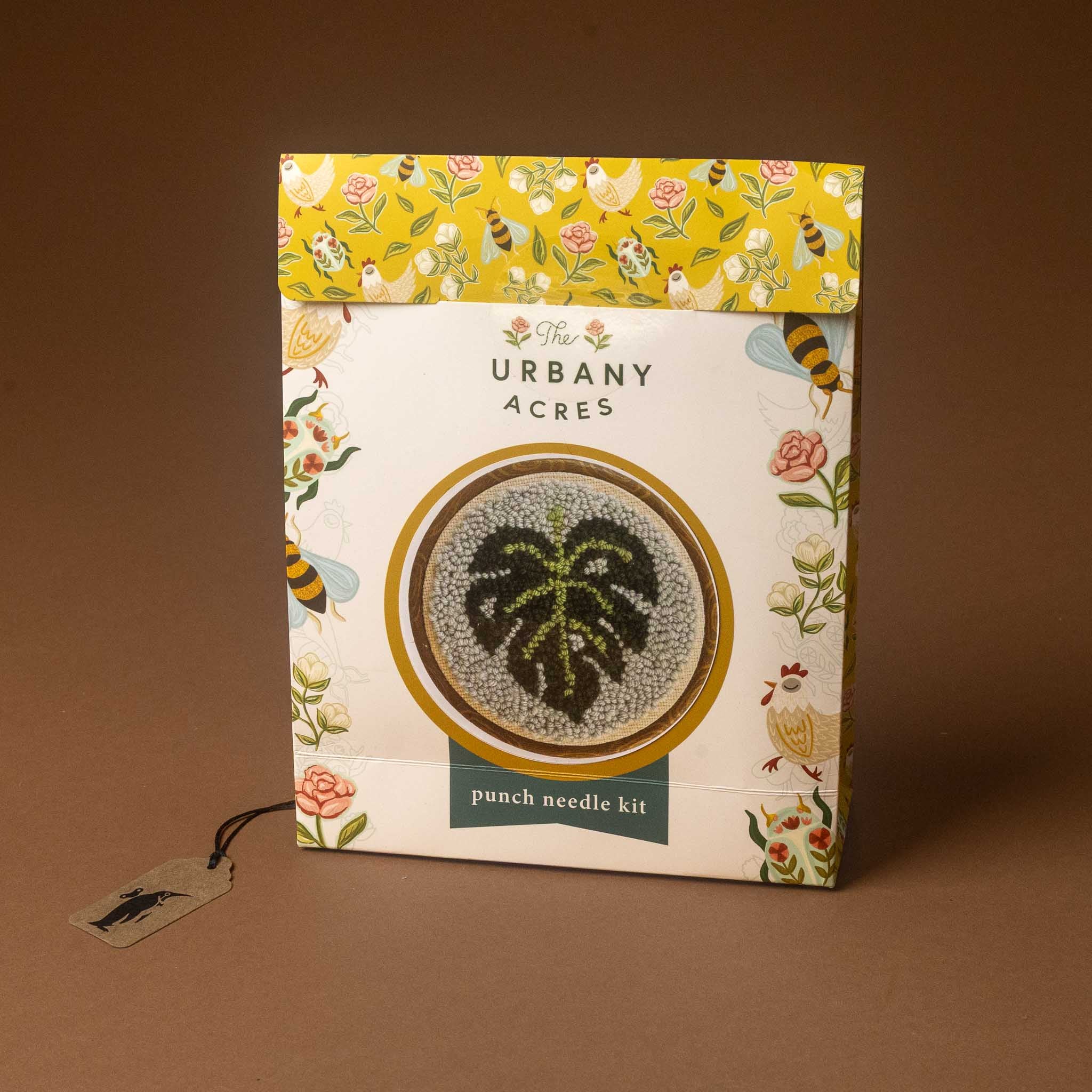 The punch needle kit box on a solid brown background. The packaging is white with a yellow floral patterned top flap, featuring a central circular graphic of a green monstera leaf on a gray and gold background.