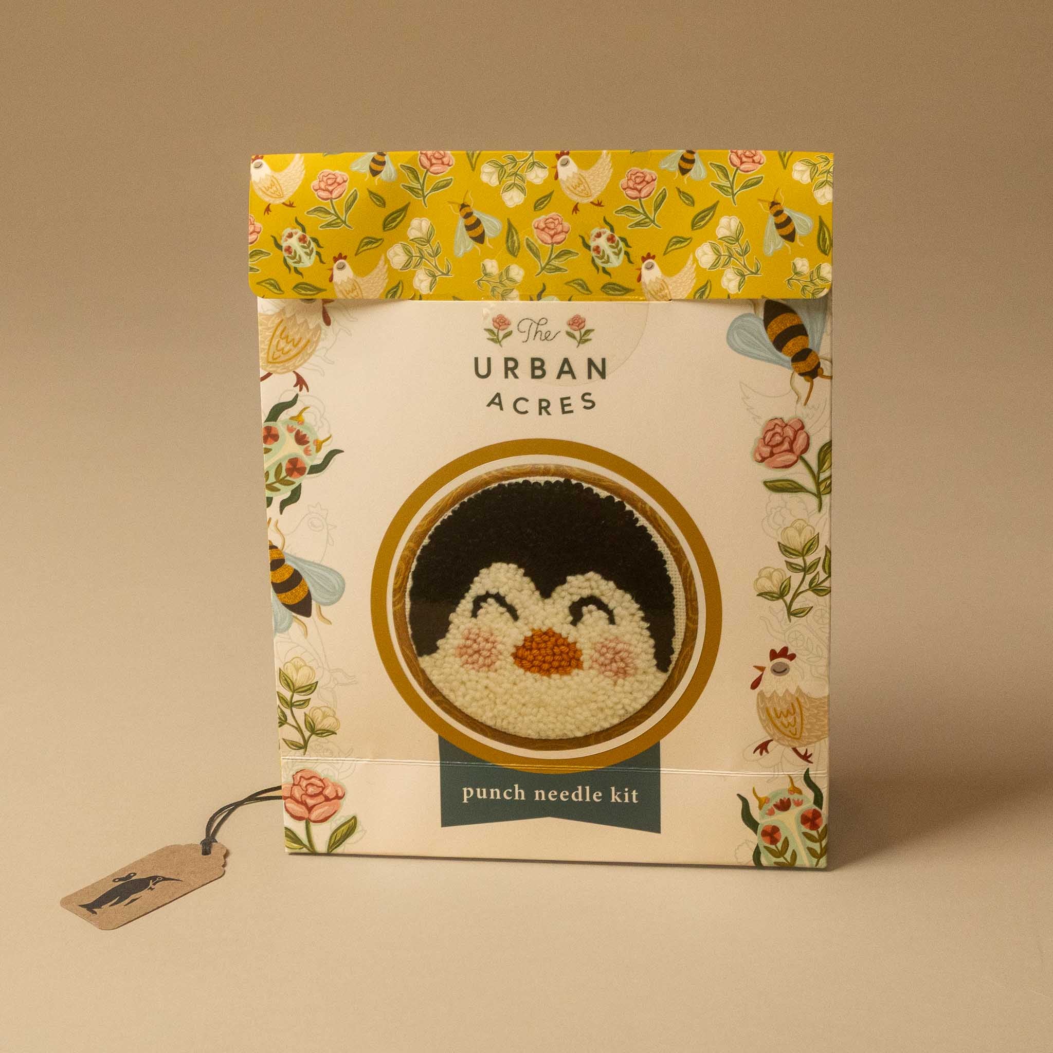 The punch needle kit in its illustrated packaging, featuring a penguin design on the front cover with a yellow floral patterned top flap and a hanging paper tag.