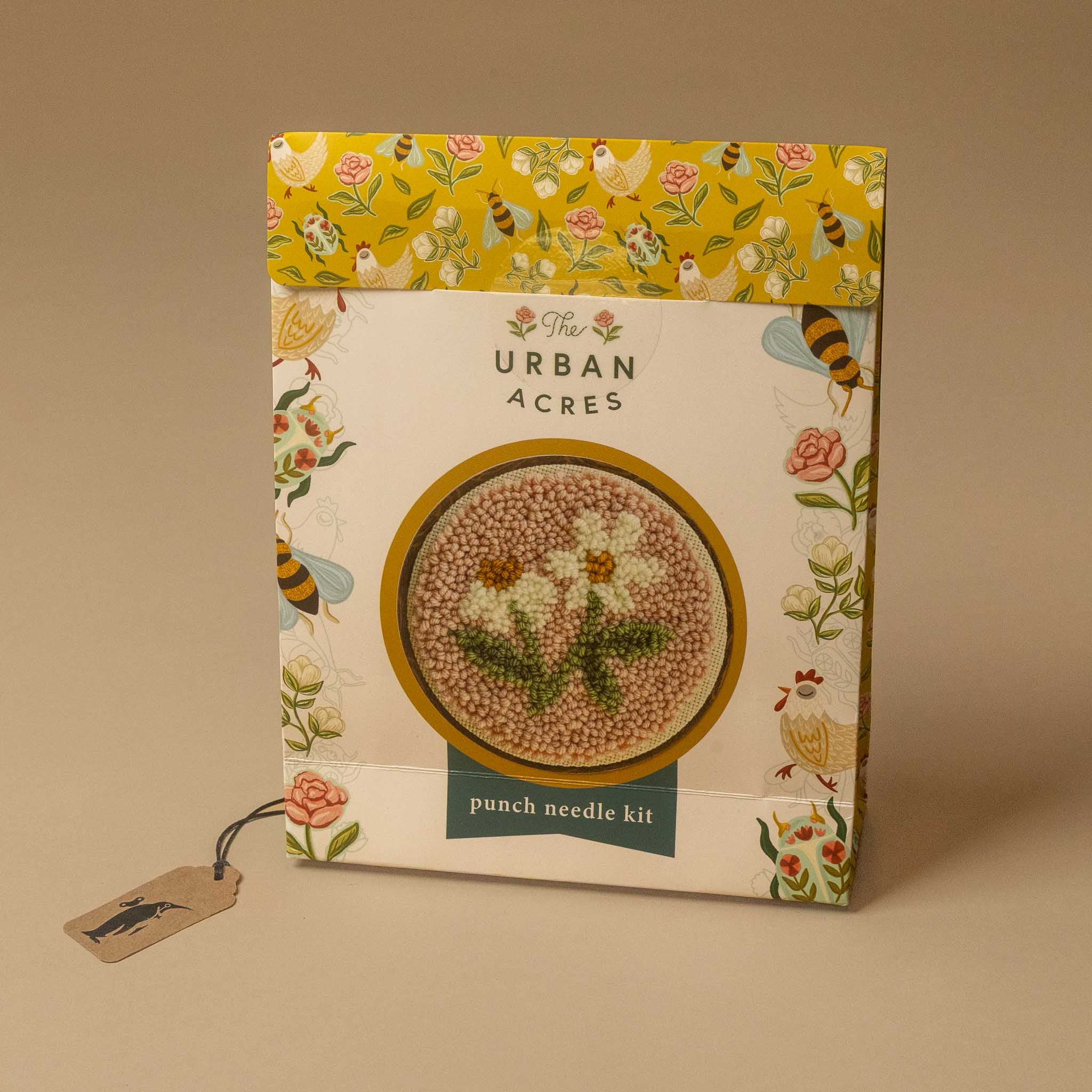The punch needle kit box on a warm beige background. The packaging features a yellow patterned top band with birds and flowers, and a central white panel with a circular cutout showing a floral punch needle design.