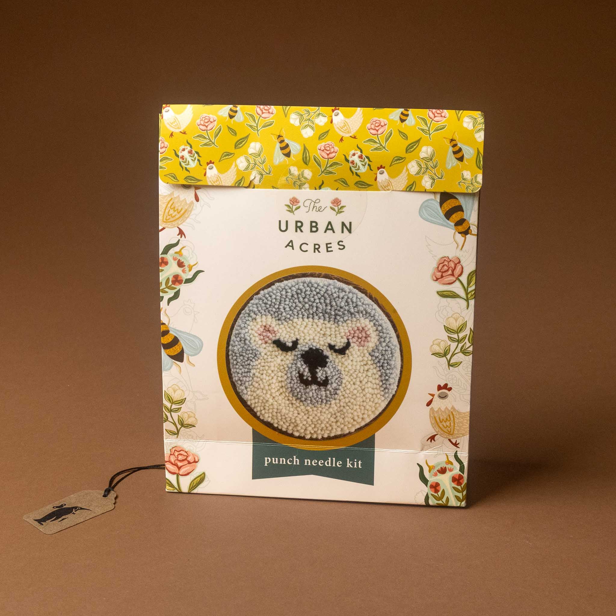 The punch needle kit in its original box, featuring a white box with a yellow floral patterned top flap and a central circular preview of a bear face. A small hanging tag is visible on the left, set against a solid brown background.