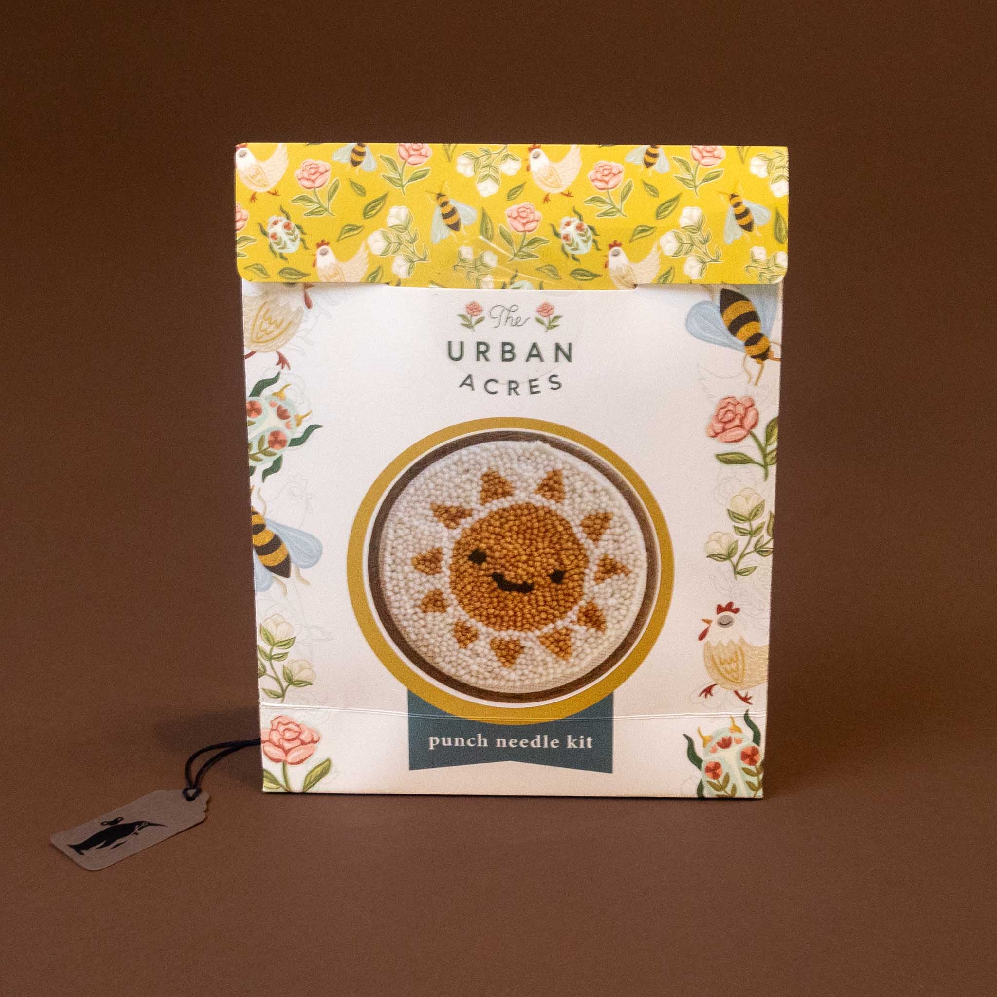 punch-needle-kit-sunshine-y-day in its original packaging, featuring a white box with a yellow floral patterned top flap and a central illustration of a sun made of punch needles. The box is displayed on a solid brown background with a small product tag hanging from the side.