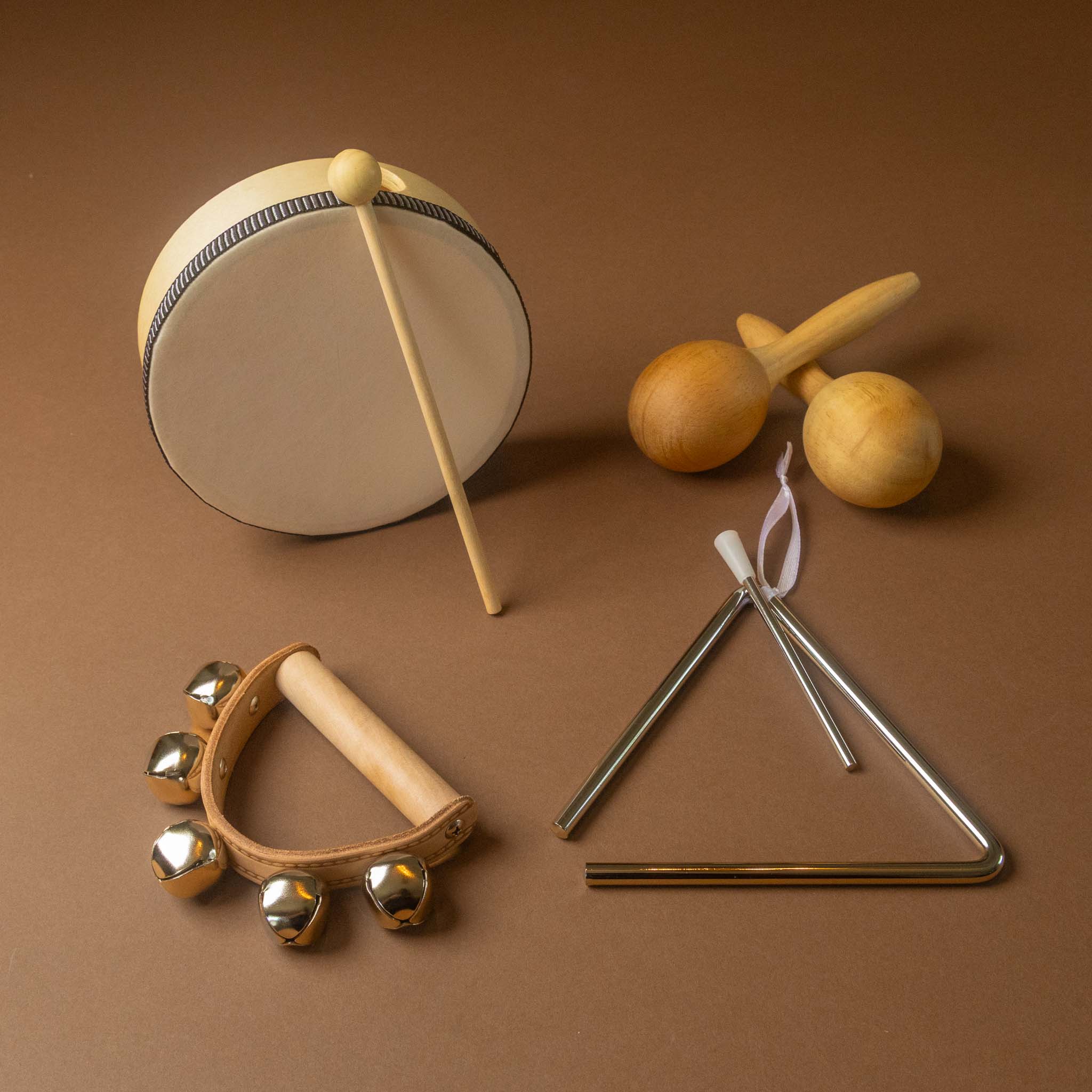 quartet-set-of-music of wooden and metal baby percussion toys arranged on a solid brown background, including a drum with mallet, maracas, jingle bells in a cymbal holder, and a metal triangle with sticks.