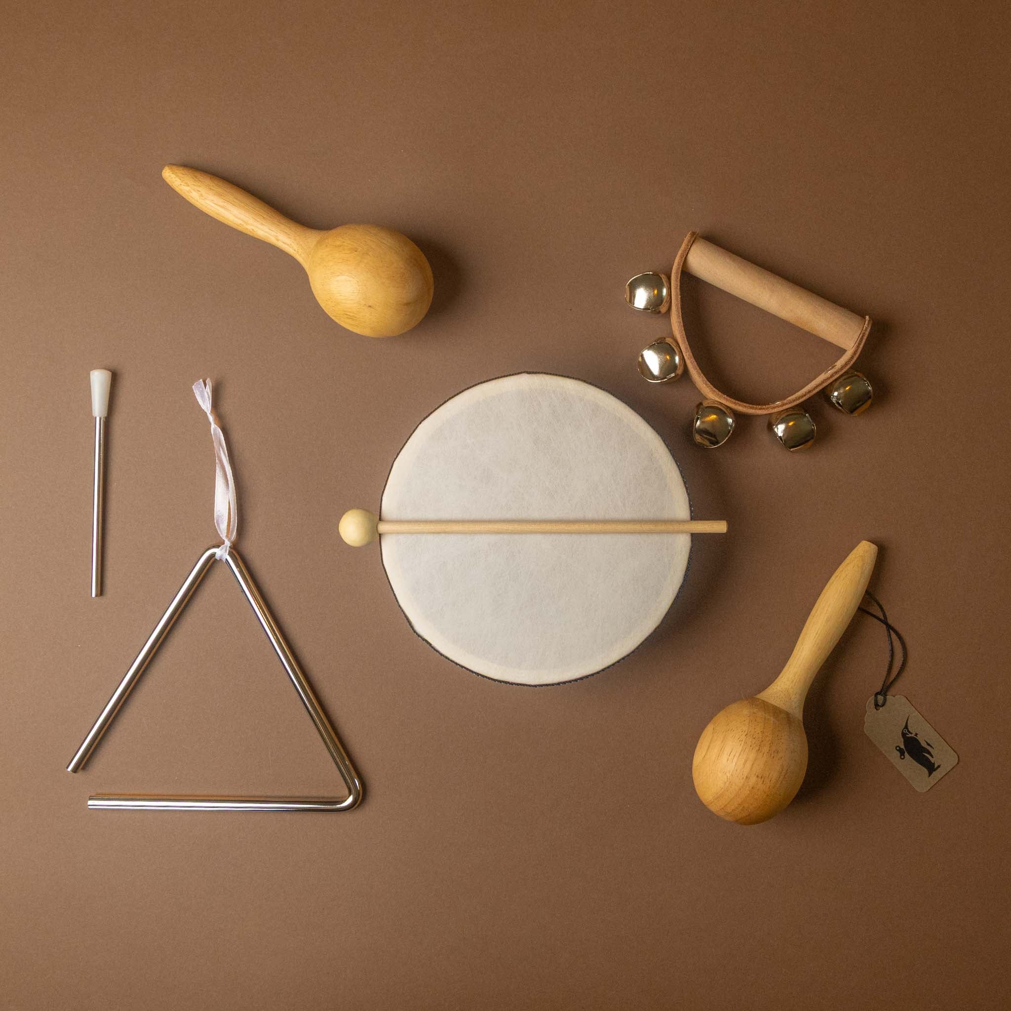 quartet-set-of-music of wooden and metal baby percussion toys arranged on a solid brown background, including a triangle, a small drum with mallet, maracas, and a jingle bell hand drum.