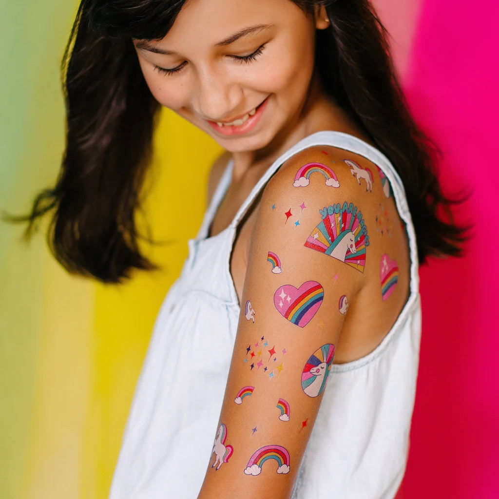 Girl with colorful trainbow-unicorns-temporary-tattoo-sheet on her arm against a multicolored background
